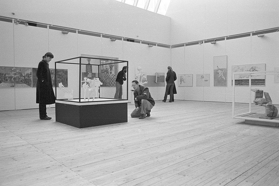Bild inifrån utställningshallen. På golvet står två kub-strukturer med skulpturer i. Fyra personer befinner sig i rummet och kollar på konsten.