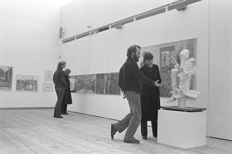 Inifrån utställningshallen. Till höger i bild står en piedestal på golvet med en vit skulptur på. Två personer står bredvid och tittar på den. På väggarna sitter det tavlor och längre bak i rummet står två personer och tittar på dessa.