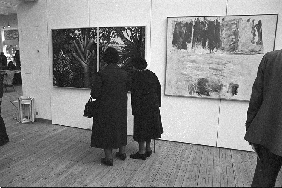 Två äldre damer fotograferade snett bakifrån samtidigt som de tittar på en större tavla som sitter uppe på väggen. Bredvid den till höger syns en annan tavla i samma storlek.