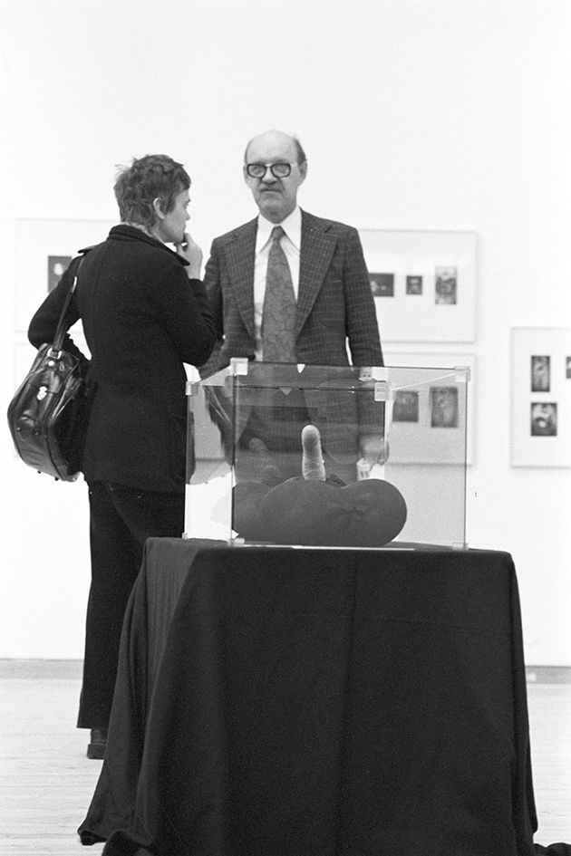 I förgrunden står en svart piedestal med låda i glas på. I lådan finns ett konstverk. Bakom konstverket står två personer varav en kollar in i kameran och den andra är vänd från kameran.
