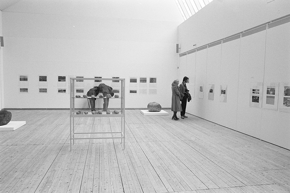 Utställningshallen fotograferad med tavlor på väggarna. Mitt i rummet står ett glasskåp. I två hörn av rummet står stora stenar på plattor på golvet. Fyra besökare står i två grupper om två och tittar på konsten.