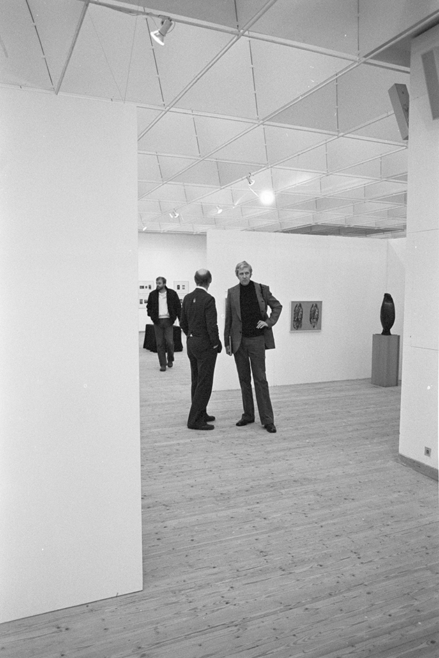 Fotografi inifrån utställningshallen. Centrerade i bild syns två män. En man tittar mot kameran, den andra har vänt huvudet mot kameran. Längre bak i rummet syns ytterligare en person.