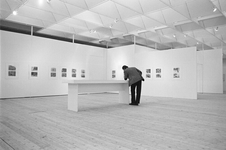 Utställningshallen fotograferad med tavlor på väggarna. Mitt i rummet står ett bord. En person står böjd över bordet och tittar på det.