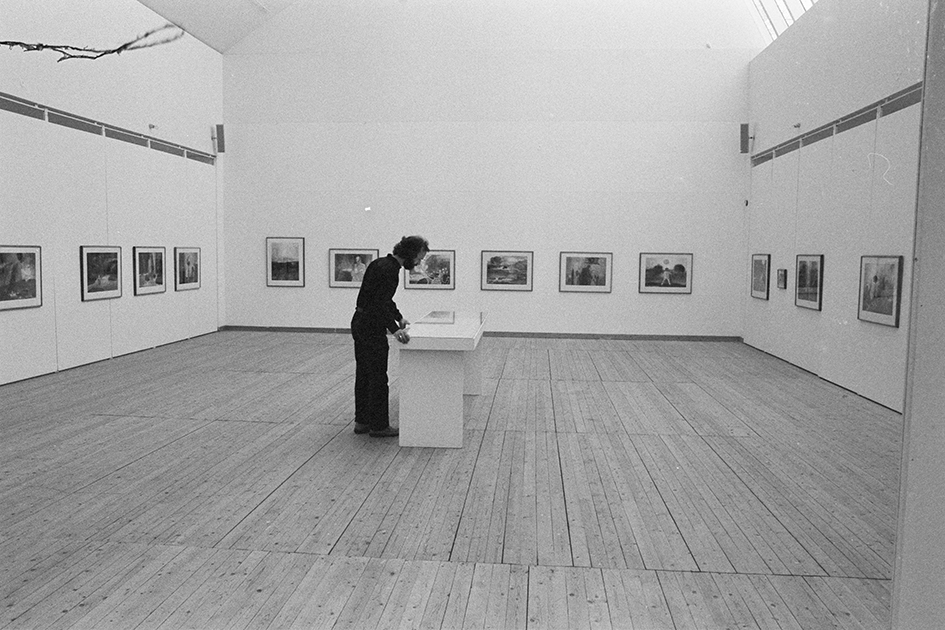 Utställningshallen fotograferad med tavlor på väggarna. Mitt i rummet står ett bord. En man står vid bordet och tittat ner på det.