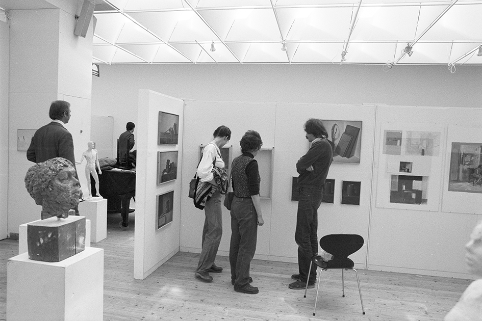 Tre personer står och tittar på ett konstverk som hänger på en vägg. Fotograferade bakifrån. Det händer fler verk på de andra väggarna och i förgrunden står en piedestal med liten skulptur av ett huvud.
