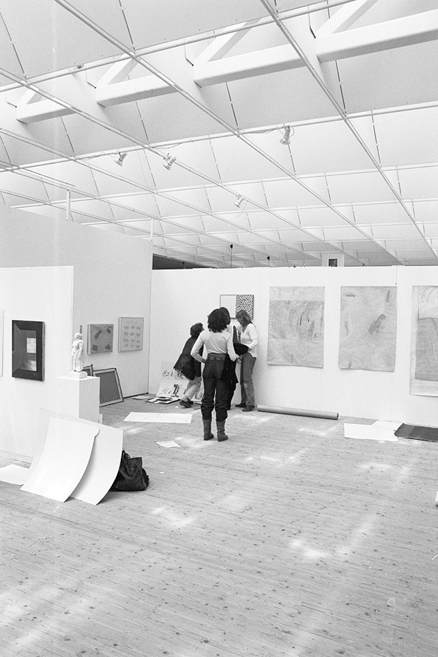 Inifrån utställningshallen. Tre personer fotograferade bakifrån står mot en vägg och sätter upp konst. På golvet ligger några papper utspritt.