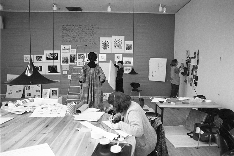 In the foreground, a person sits at a table working with paper and pen. In the background, three people are putting up art on the walls. Overall, there is a bit of a mess around.