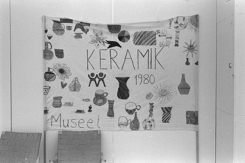 Ett tygstycke där det står "keramik 1980 museet". På tyget finns även ett antal illustrationer av olika bruksföremål och blommor.