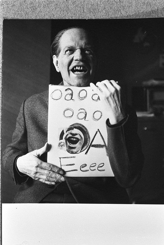 Fotografi av ett fotografi. En man håller upp ett papper. Han har munnen öppen, visar tänderna och tittar upp mot höger. På pappret står det ett antal bokstäver.