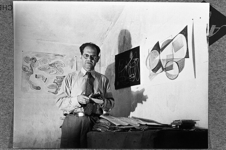 Fotografi av ett fotografi. En man klädd i skjorta och slips fotograferad tomt rum med några konstverk på väggen runt sig.