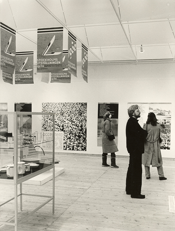 Tre besökare i utställningshallen. Till vänster i bild syns en glasmonter med olika bruksföremål. På väggen hänger stora fotografier uppsatta. I taket hänger affischer från Stockholmsutställningen 1930.