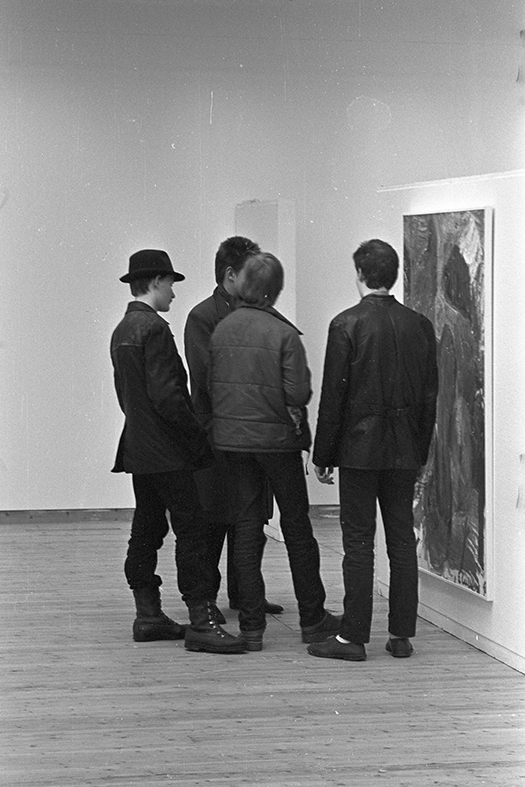 Svartvit bild. Fyra killar står framför en tavla och verkar titta på den. Två står med ryggen mot kameran, en lite från sidan och den fjärde skyms av de andra killarna.