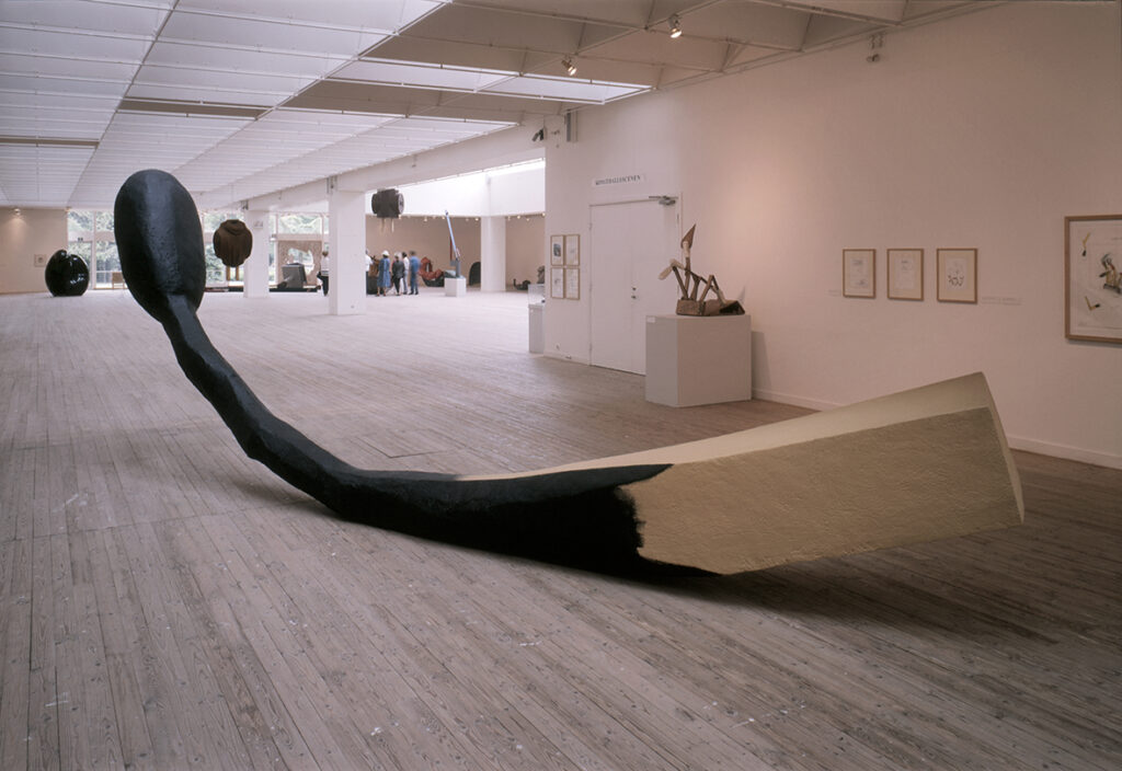 Ett konstverk. En tändsticka i jättestor storlek, som är svart halvvägs ner för stickan ligger på golvet inne i utställningshallen.
