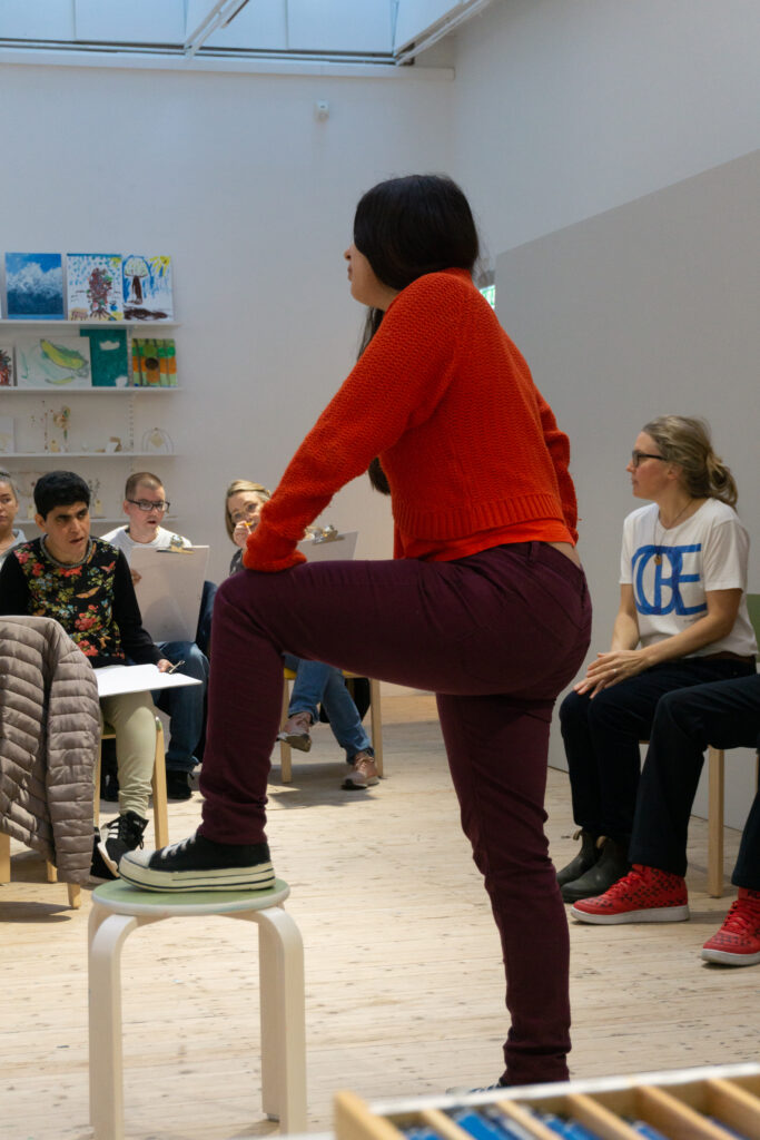 A girl in a red shirt and burgundy pants is modeling for a group of people drawing her.