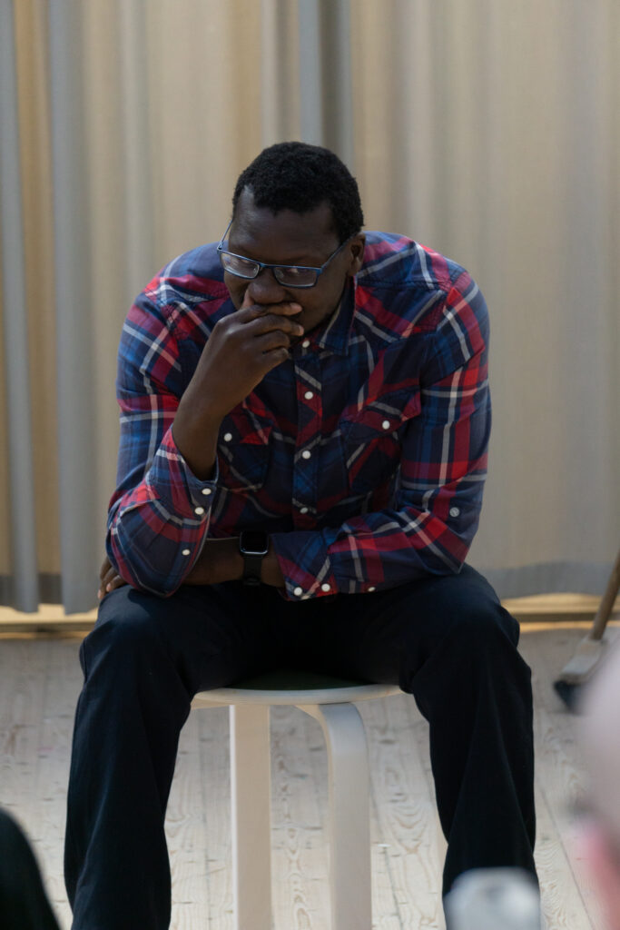 A man is sitting on a stool, slightly leaning forward with elbows on knees and wearing jeans, a checkered shirt, and glasses.