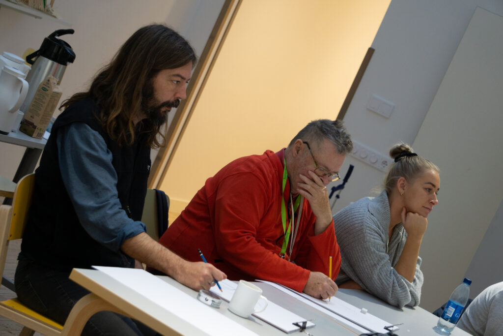 Three people are sitting at a table in the workshop drawing on large sheets of paper.