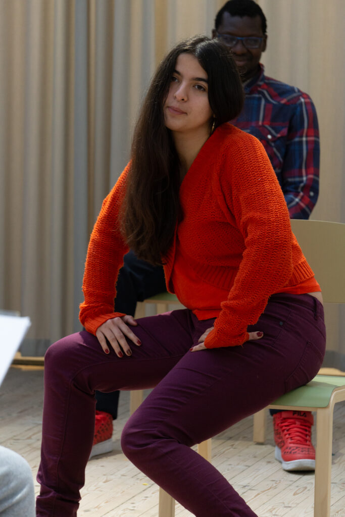 En person sitter på en stol, fotograferad framifrån. Personen lutar händerna mot sina lår och låter ena knäet sjunka ner lite.