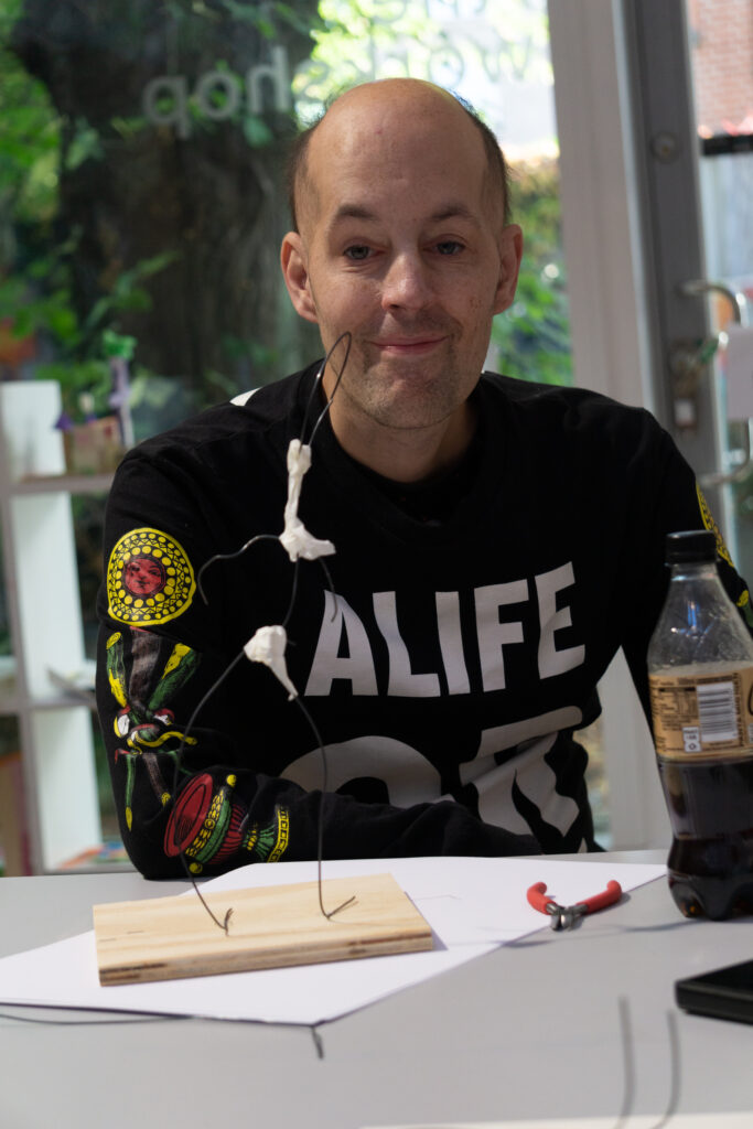 Porträttfoto av en person som sitter vid ett av borden i verkstaden. Framför personen på bordet står en liten ståltrådsskulptur och en cola-flaska.