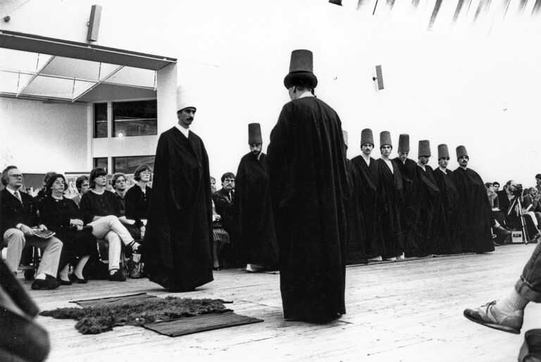 Dansade_Dervisher. En person i svart cape och hög hatt står med ryggen mot kameran, ungefär tio personer står och tittar på den första personen, iklädd liknande kläder. Publik sitter längs väggarna.