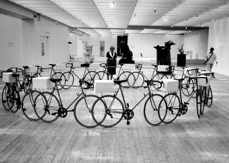 Ett konstverk. Ett tiotal cyklar står i ihopsatta i en stor ring inne i utställningshallen. Bakom denna ring ses en besökare titta på nyss nämnda konstverk.