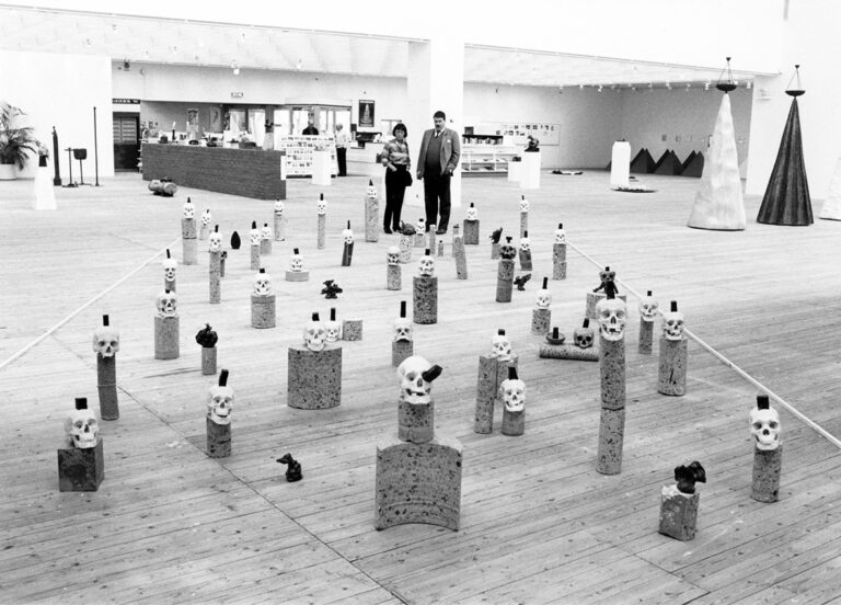 Många dödskallar med olika former placerade på huvudet. Ett 30tal står utställda i utställningshallen. Två personer står i bakgrunden och tittar på verket.