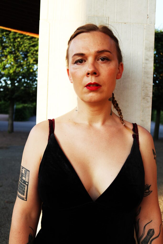 Artisten Maja Darkest Karlsson. Hon är klädd i svart och har tatuerade armar och rött läppstift.