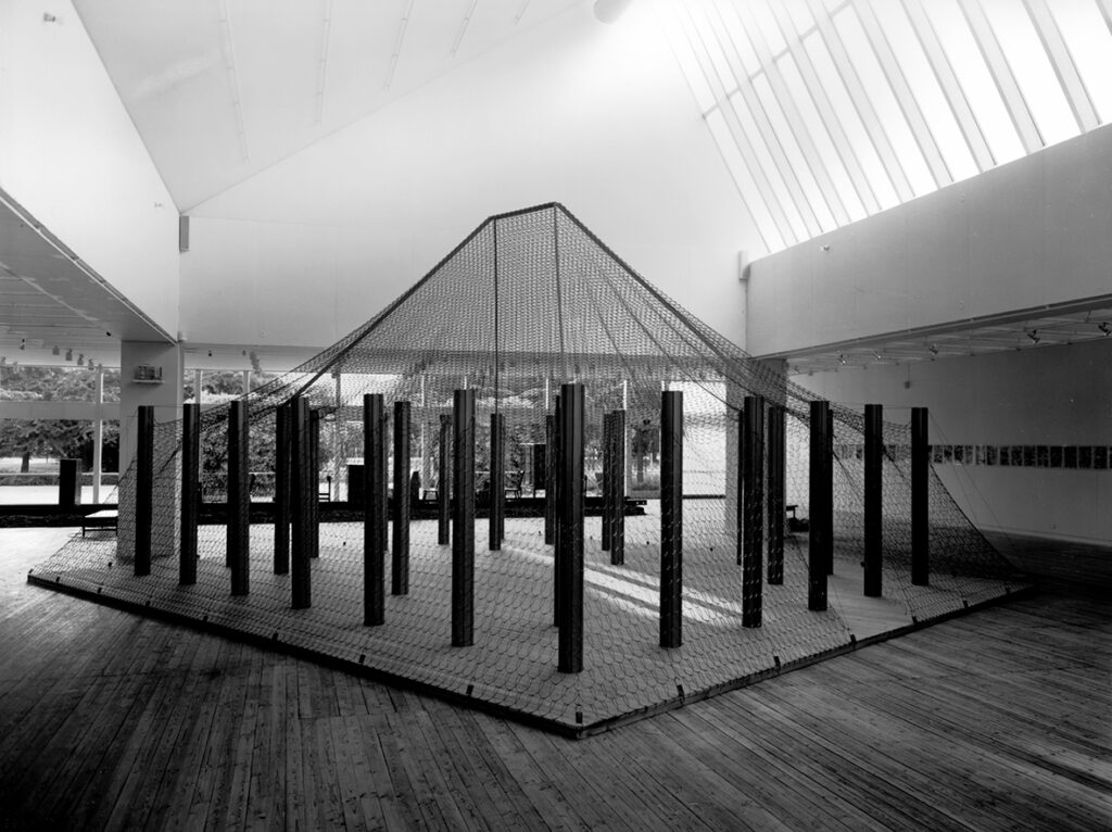 Ett konstverk i utställningshallen. Konstverket består av en stor platta där det står ett antal stolpar. Plattan och stolparna är under ett stort nät.
