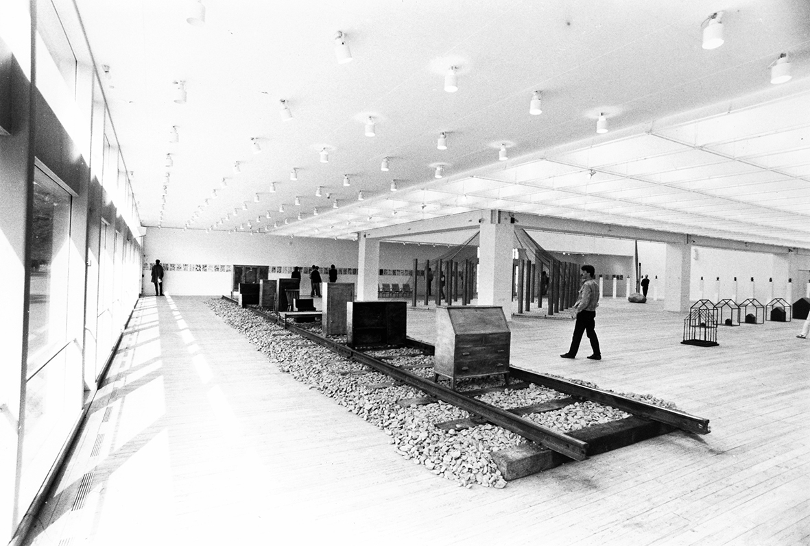 En bild inifrån utställningshallen. Närmast kameran syns en stor tågräls när ett tiotal byråer står utställda. Runtom i hallen ser man andra konstverk samt besökare.
