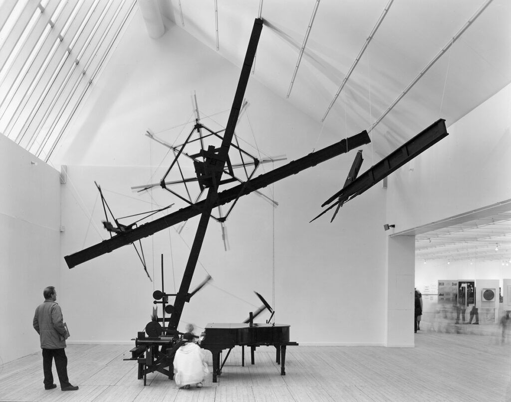 Konstverk i utställningshallen.En större mörk konstruktion med olika stolpar och former, samt ett piano.