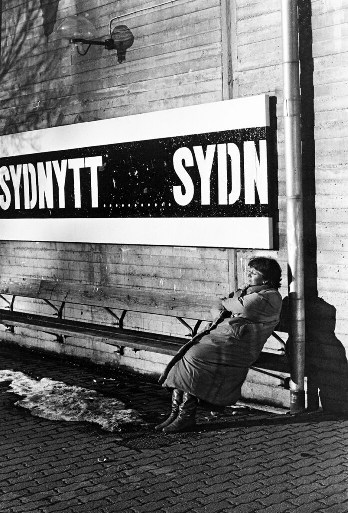 En kvinna iförd en vinterjacka sitter på en bänk utanför Malmö Konsthall. Bakom kvinnan syns en stor skylt med ordet 