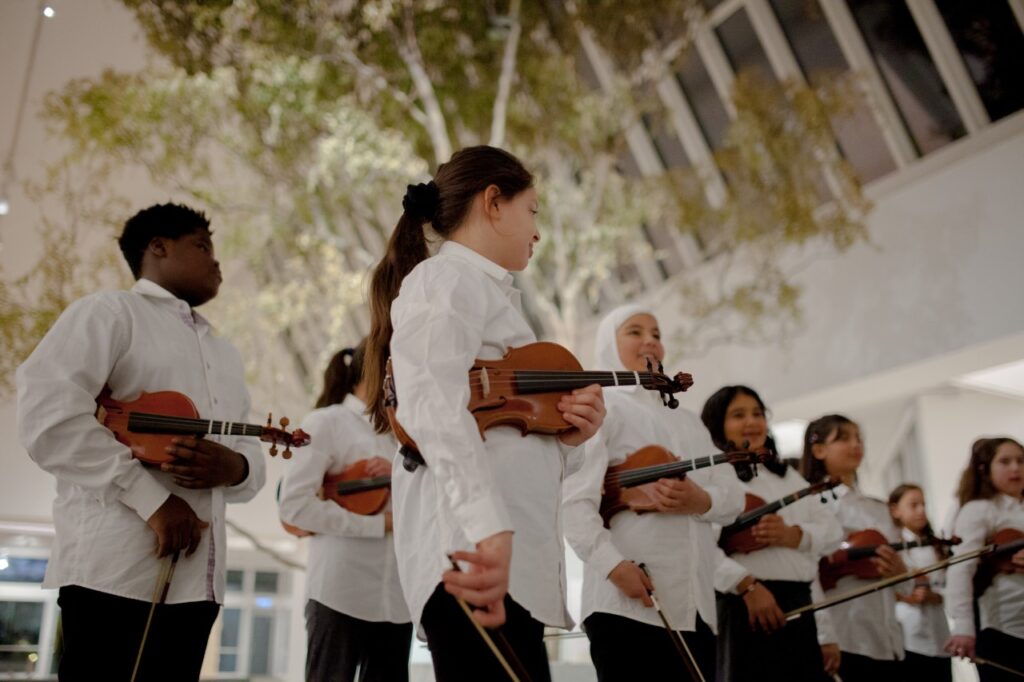 En elevorkester, cirka tio barn syns på bild. De har vita skjortor och fioler och tvärflöjter i händerna.