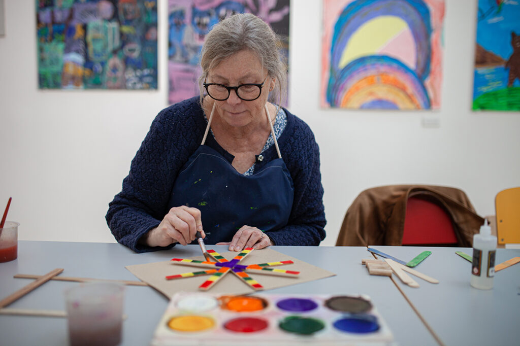 Äldre kvinna målar koncentrerat i en färgglad verkstad.