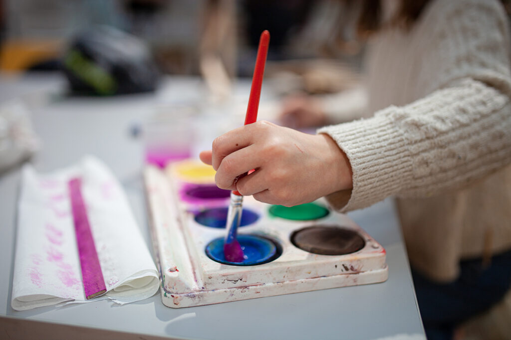 A child's hand holds a paintbrush and dips it into a palette of colors