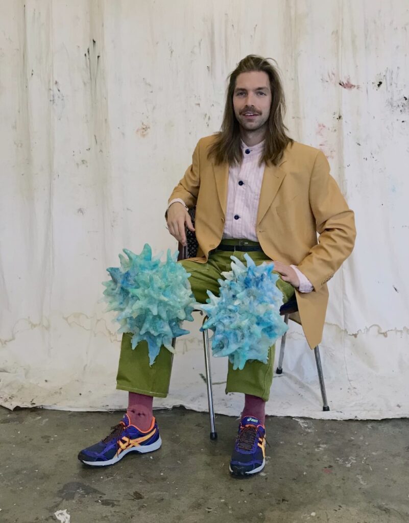 Porträtt av yngre man som sitter ned i klädd i glada färger med organiska objekt på sina knän