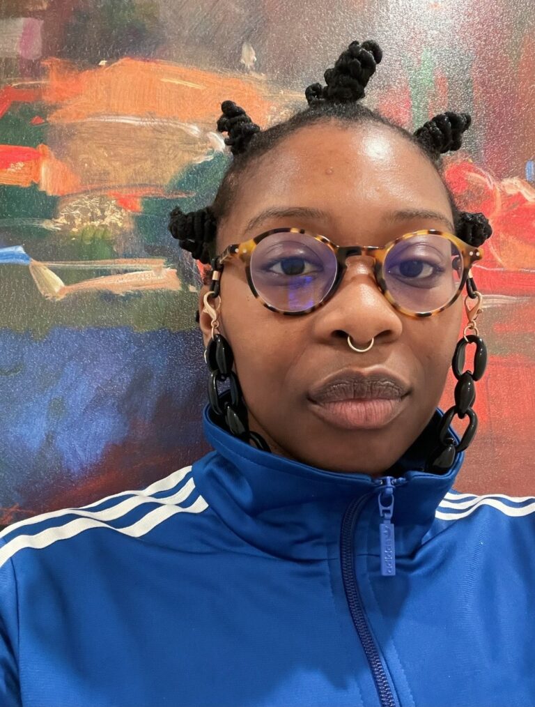Porträtt/selfia Zoé Samudzi i blå adidaströja och mot en färgglad bakgrund.
