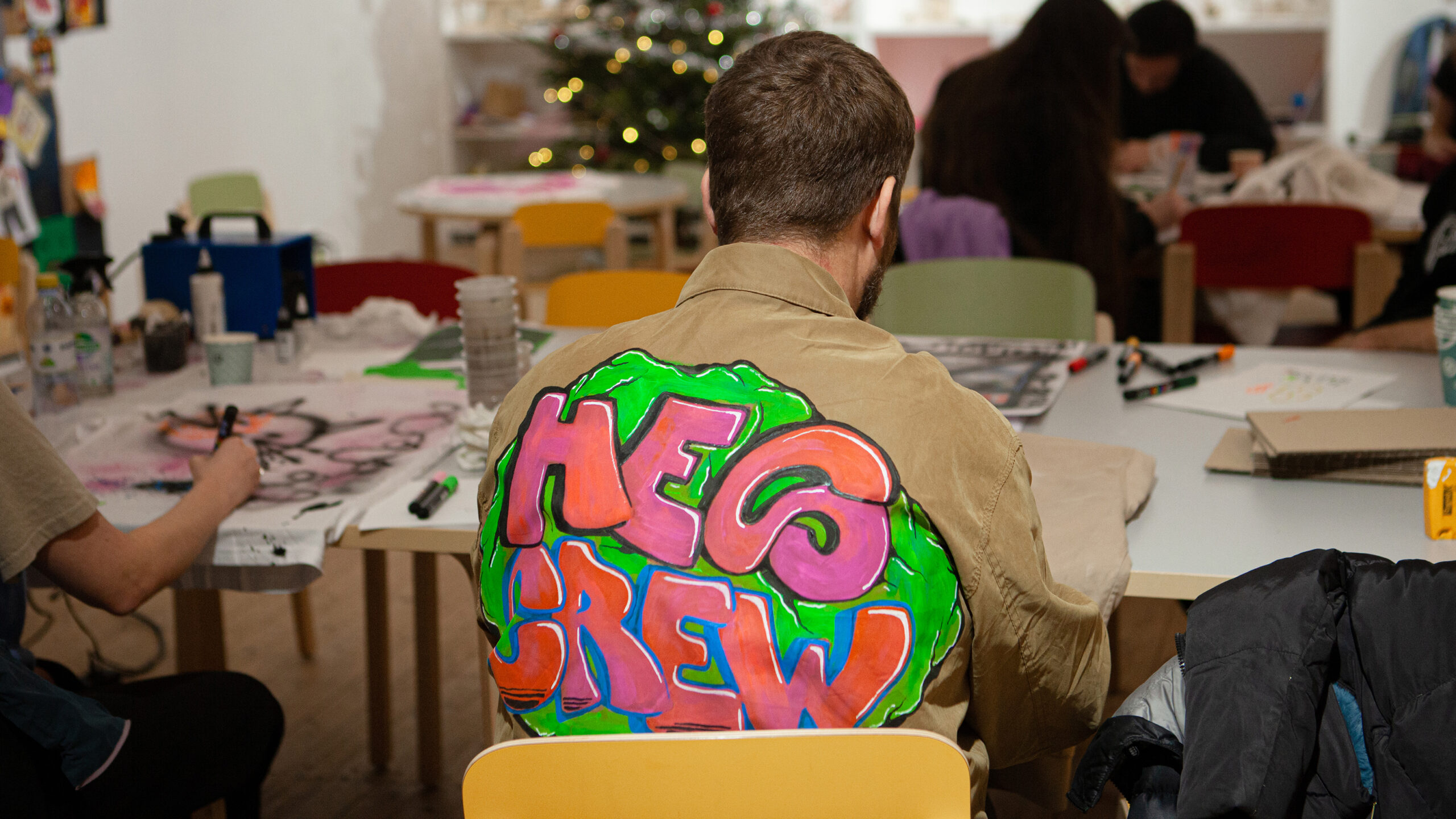 Artist Ossian Theselius from behind, sitting during his workshop on the theme of 'Back Pieces.' He wears a beige jacket with a colorful graffiti design on the back.