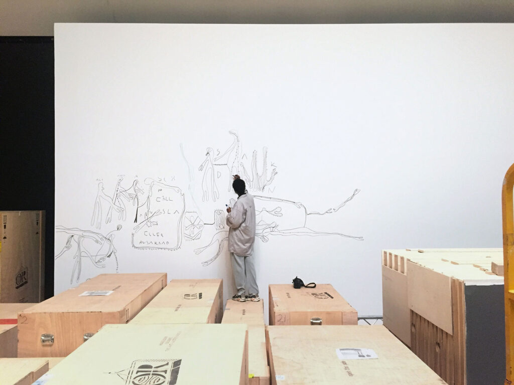 Bild av konstnären Fatima Moallim när hon teckar en stor teckning på vit vägg i vad som ser ut att vara en lagerlokal.