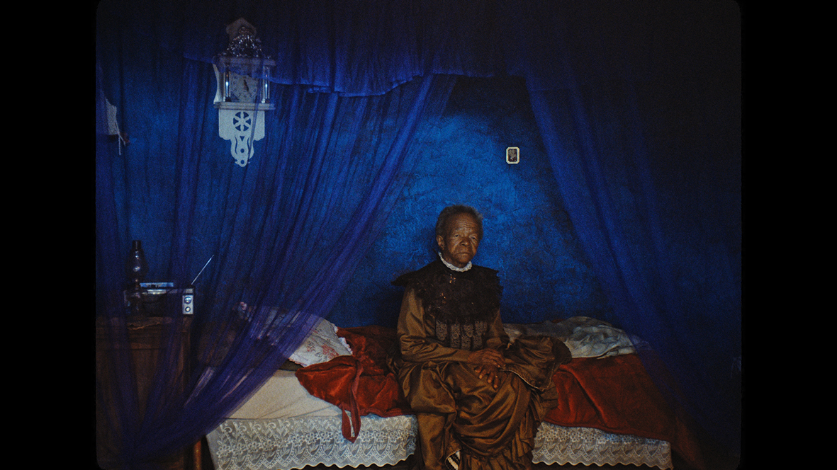 En äldre kvinna sitter på ett rött täcke i en säng med blå sänghimmel.