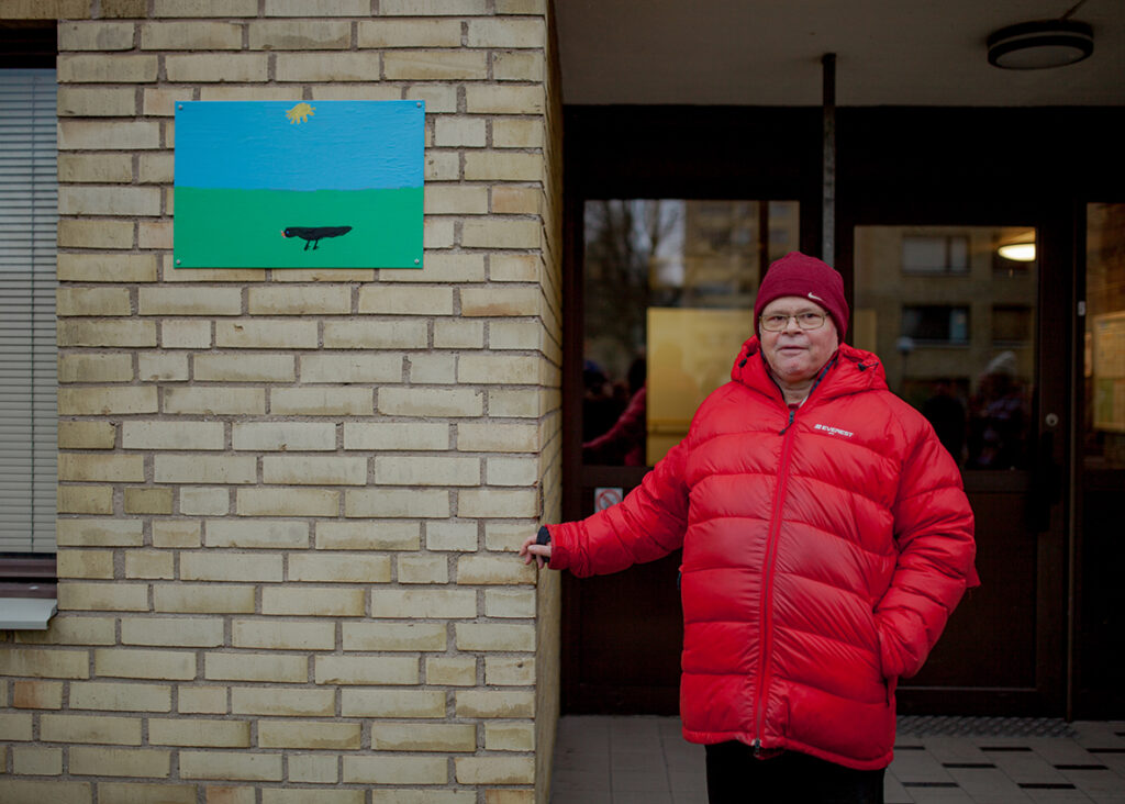 A man stands in front of a house wall wearing a red jacket and hat. He stretches his arm towards a piece of art on the house wall.