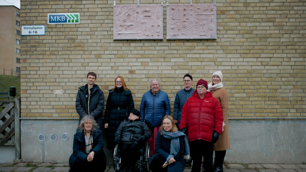 Grupp människor står framför en gul husvägg med två konstverk på väggen ovanför sig.