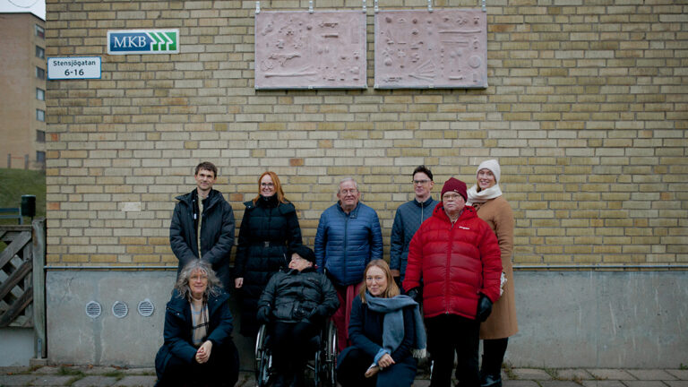 A group of people standing in front of a yellow house wall with two artworks displayed above them.