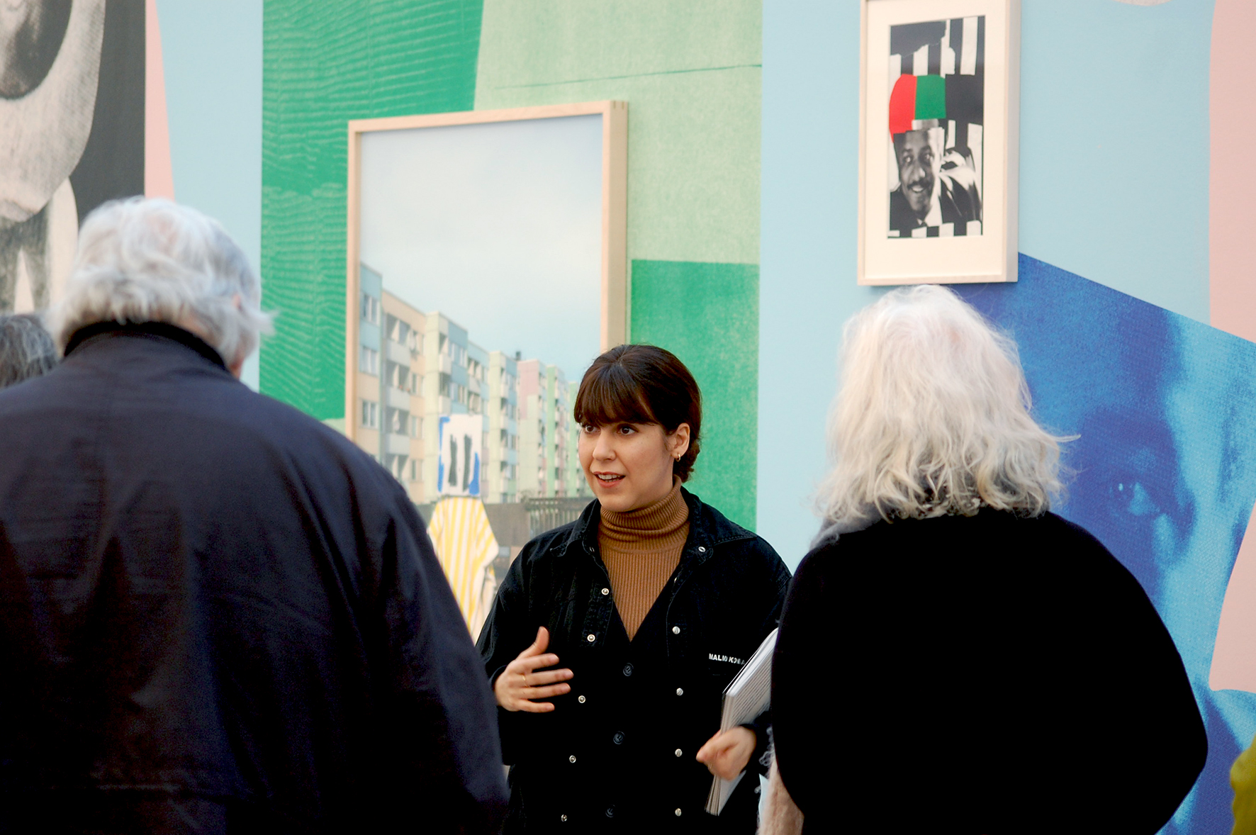 One of the art gallery guides narrates an exhibition. In addition to her, two visitors are portrayed with their backs to the camera.