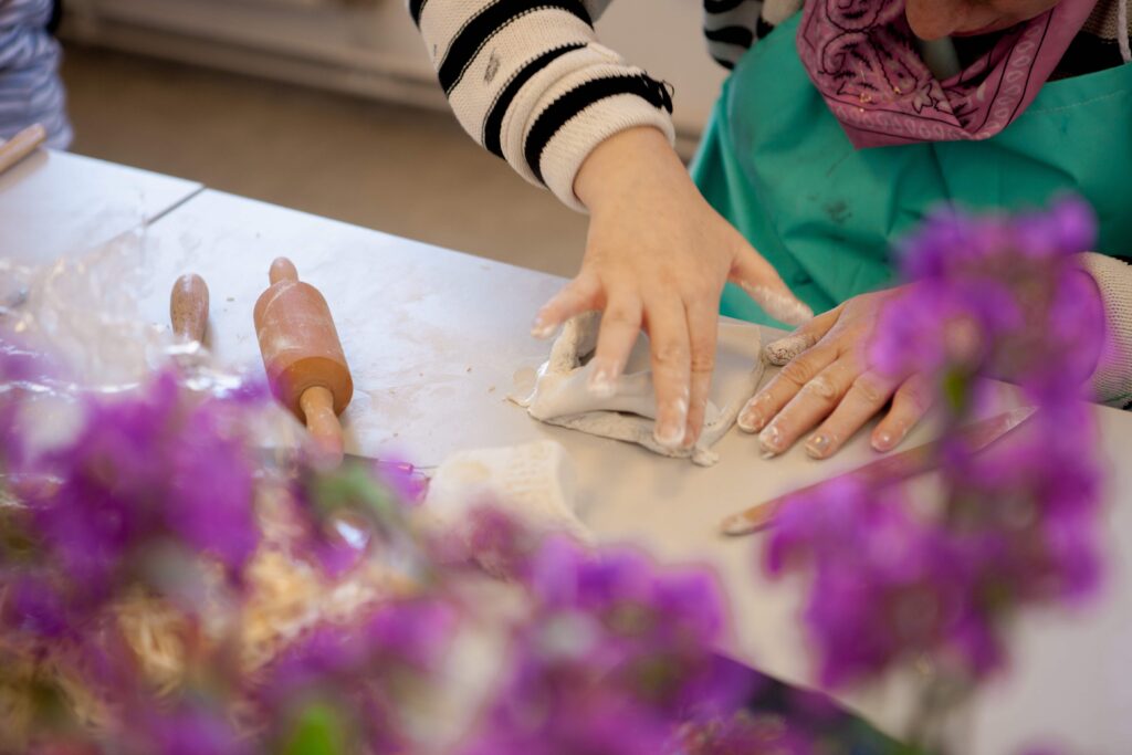 A person working with clay in the background - partially obscured by purple flowers in the foreground.