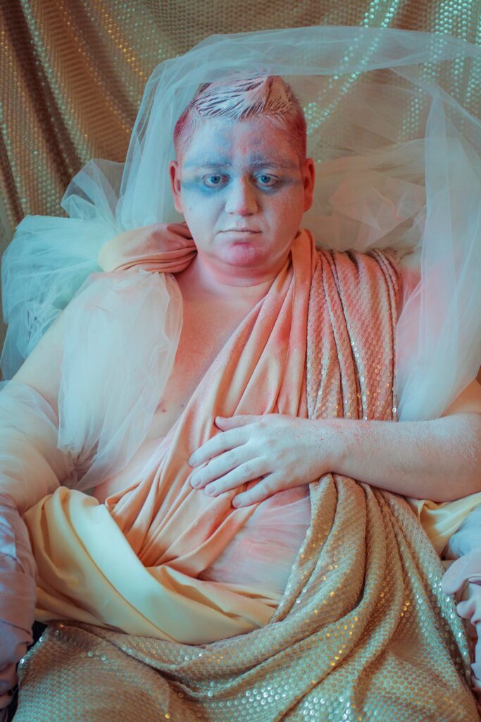 Fotografiskt porträtt av konstnären Leif Holmstrand som en Buddha-figur, färgad i rosa, guld och beige.