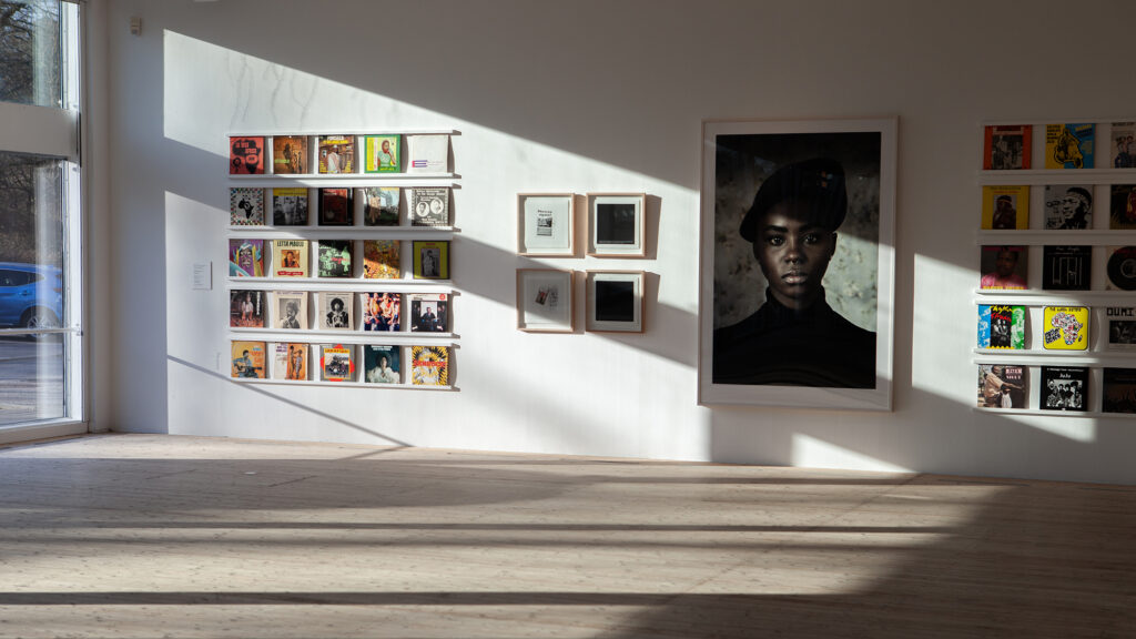 Ljuset faller vackert in på en vägg med konstverk i konsthallen; skivomslag och foton.