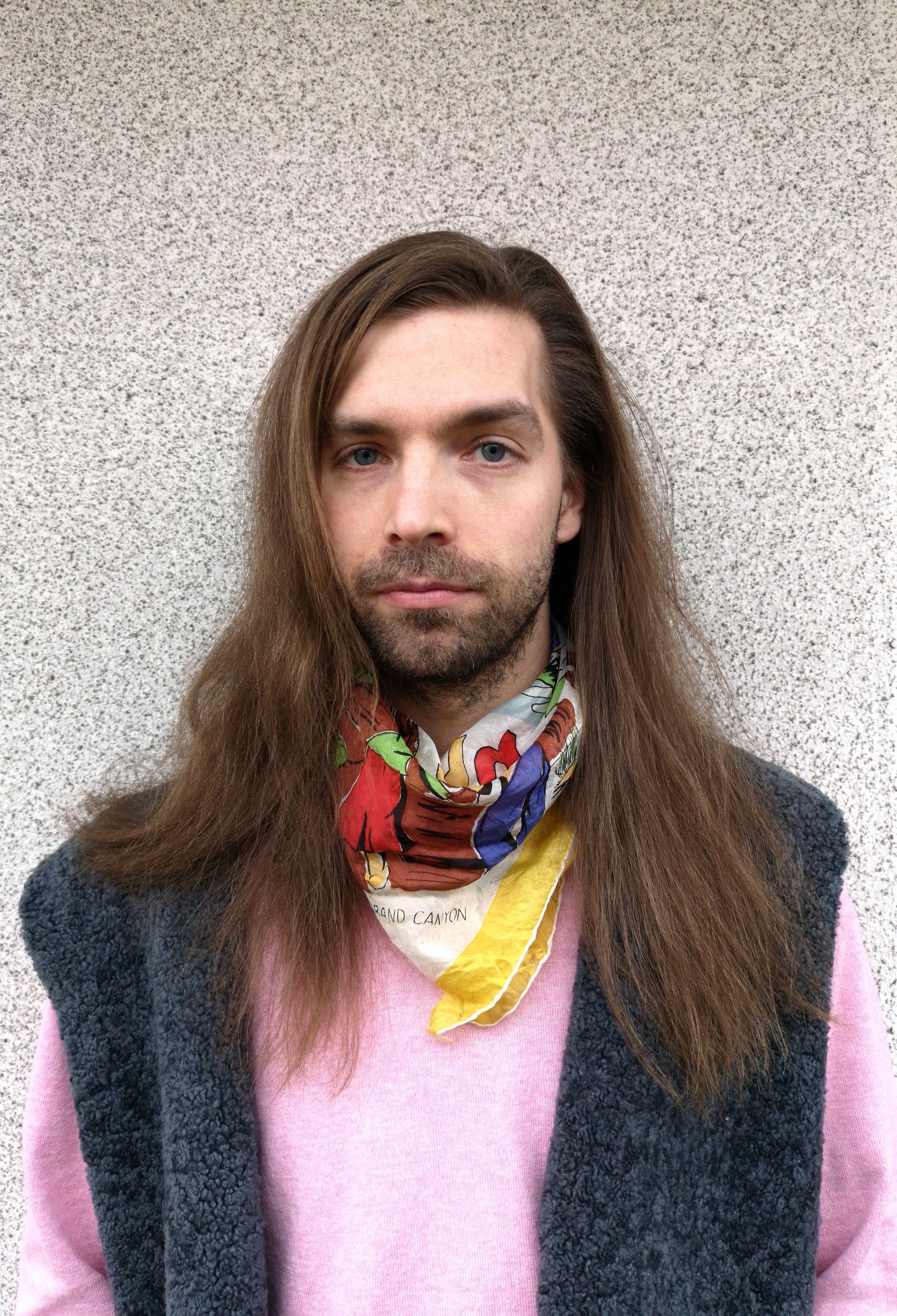Konstnären Matti Sumari, porträtt. Långt, brunt hår och iklädd en rosa tröja med väst och en gul scarf runt halsen.