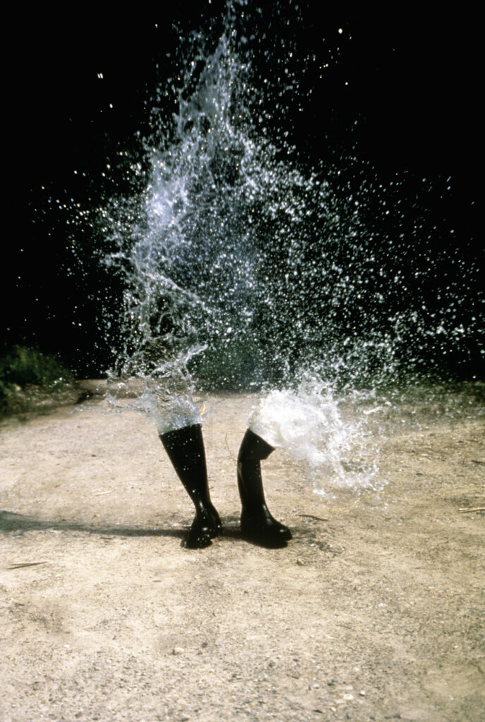 Water spraying from a pair of rubber boots standing on sandy ground.