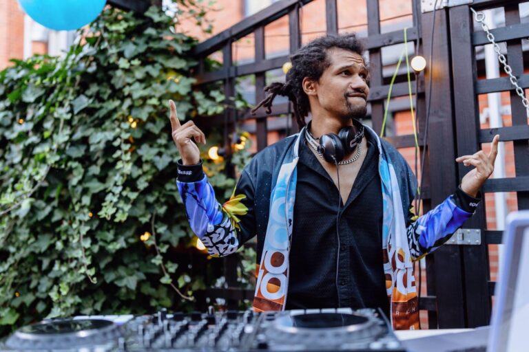 En cirka 30-årig glad man i rastaflätor står vid ett mixerbord utomhus och spelar musik.