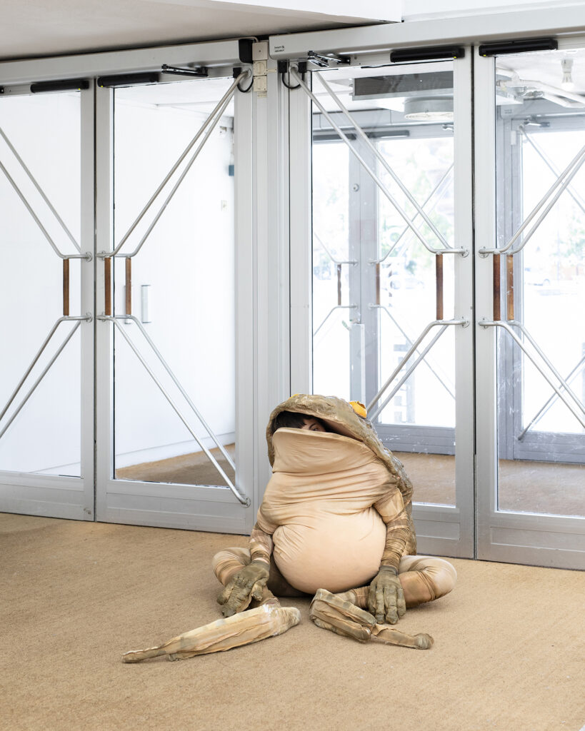 A person in a frog costume sits on the floor inside Malmö Konsthall. 