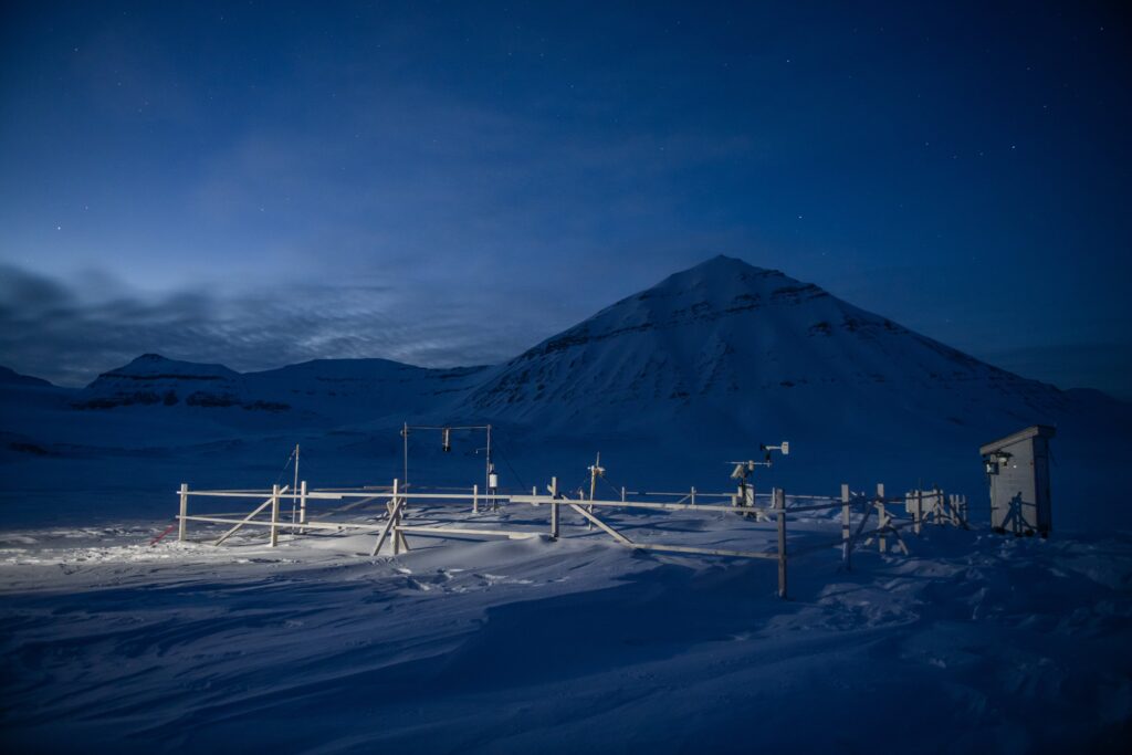 An image of a research station with various measuring instruments in the Arctic during dusk. In the background, a snow-covered mountain.